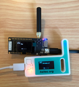 Two development boards with antennas and LCD screens sitting on a table, each connected to a microUSB power source. The LilyGO board on the left displays the message "Sup" from a user named "IFK." The Heltec board on the right displays the message "Not much, u?" from user "Js2."