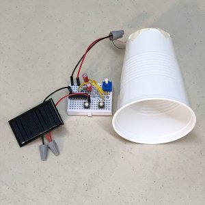 Closeup of an assembled solar synth kit: a breadboard with an integrated circuit, two switches, two capacitors, two resistors, a potentiometer, four jump wires, a solar panel, and a piezo speaker taped to a plastic cup, which acts as a horn loudspeaker.