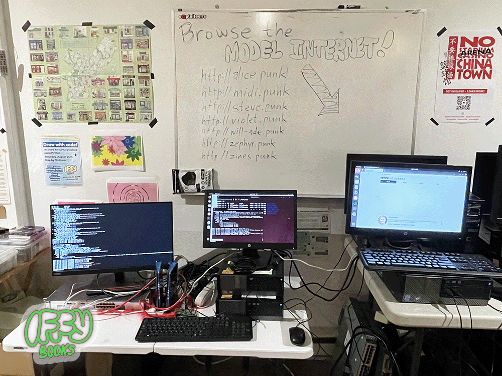 A photo of the Model Internet at Iffy Books, comprising two desktop computers, a switch, and a Raspberry Pi on a small table with two monitors. Another computer is displaying a website on the network. A white board in the background reads "Browse the Model Internet!" followed by a list of local URLs such as "http://alice.punk" and "http://zephyr.punk"