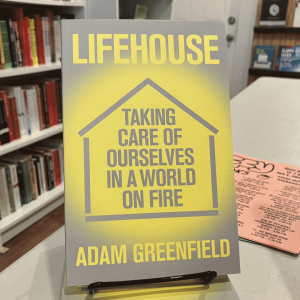 A photo of the book 'Lifehouse: Taking Care of Ourselves in a World on Fire' by Adam Greenfield, with a yellow-on-gray stencil design of a house shape surrounding the title.