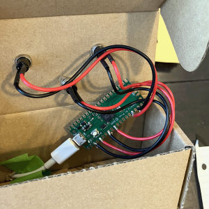 Photo of a Pi Pico microcontroller with three sets of black and white wires soldered to the pin holes. The wires connect to push button switches in the top of a cardboard box.