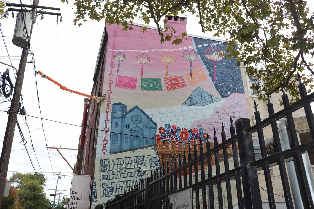 Photo of the Da Vinci Art Alliance in South Philly, with a mural that includes paper lanterns, flag decorations, and a diagram of a molecule