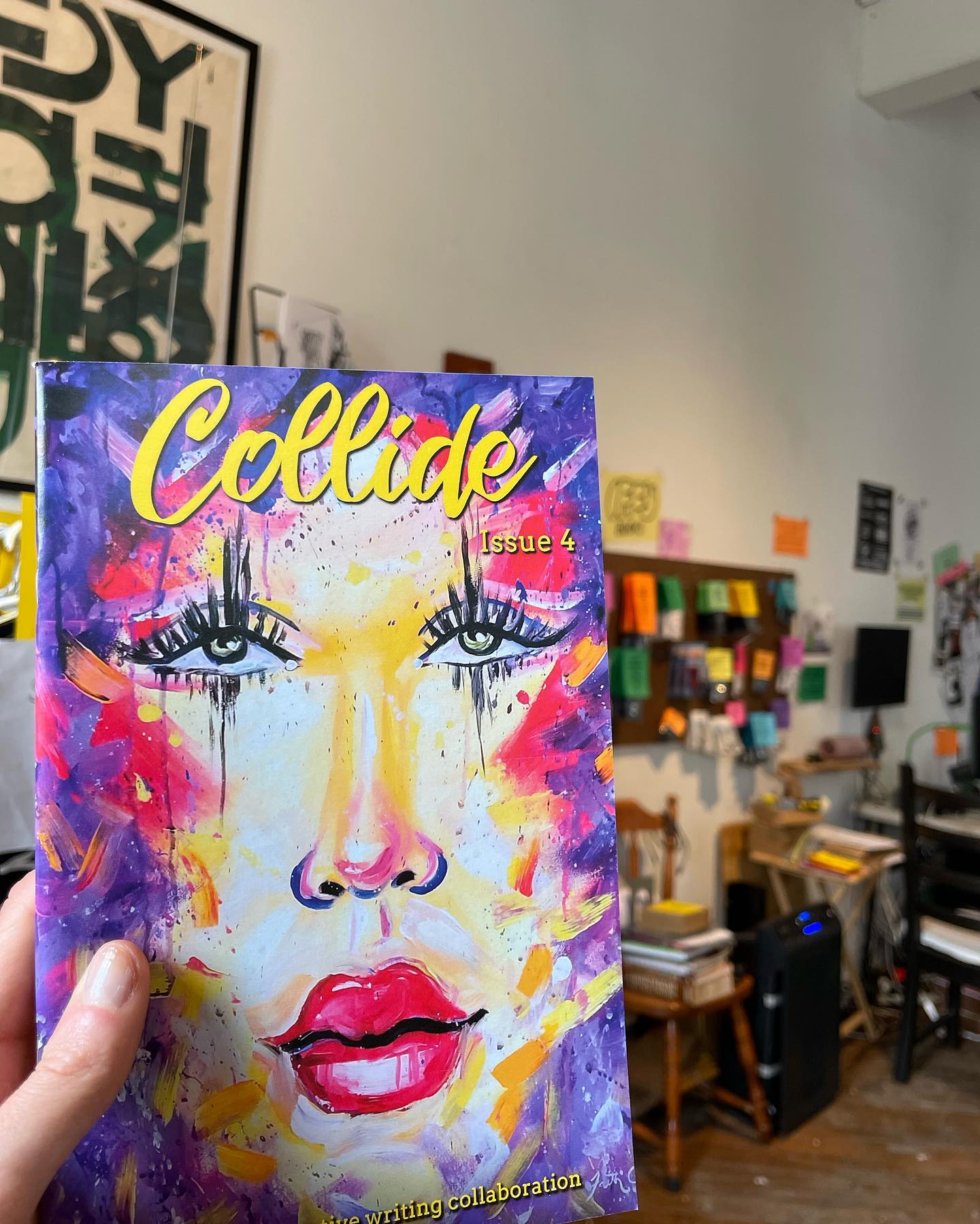 A hand holding up Collide Zine issue 4 at Iffy Books. The cover has a painting of a woman's face with bright red lips and red and purple splotches around the edges.