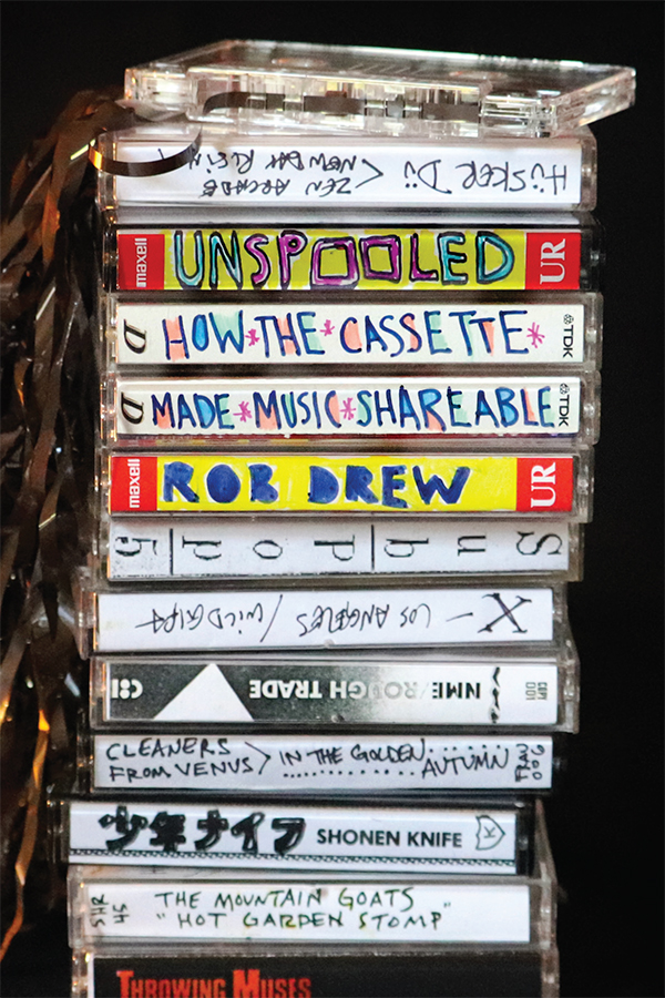 Cover of the book 'Unspooled: How the Cassette Made Music Shareable' by Rob Drew, with the title and author's name handwritten on the spines of cassette tapes, mixed into a stack of other cassettes