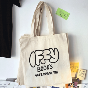 A natural cotton tote bag hanging on wall above a community flyer board. The bag has the Iffy Books logo and "404 S. 20th St., PHL" screen printed in black.