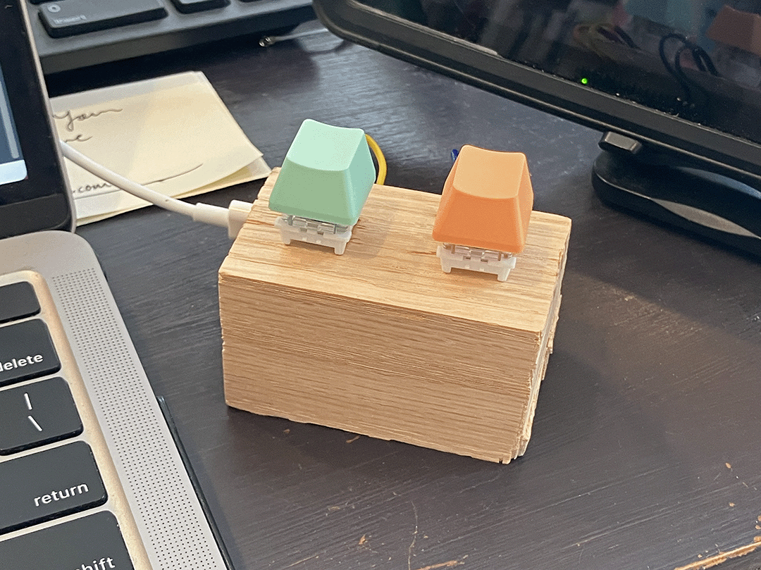 Photo of a DIY macro keyboard sitting next to a laptop: a block of oak wood with two mechanical keyboard switches mounted on top, connected to a Raspberry Pi Pico device on the rear side. The key caps are mint green and pastel orange.