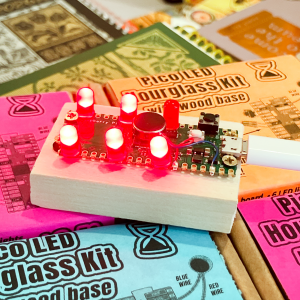 An assembled Iffy Books Pico LED Hourglass kit, with 5 out of 6 LEDs on a Pi Pico board lit up. The board is screwed to a piece of poplar wood.