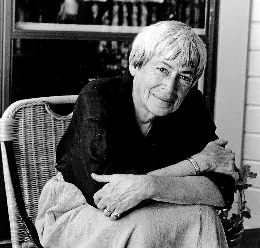 Black and white photo of the author Ursula K. Le Guin leaning forward in a wicker chair and smiling